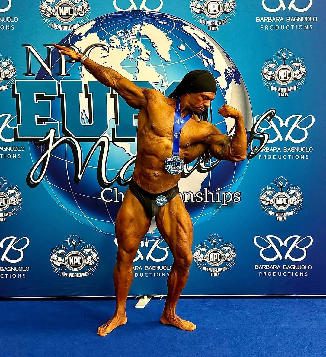 A ProFlow Physiques client alone on a competition stage, holding a side pose in front of a championship backdrop, medal round the neck.