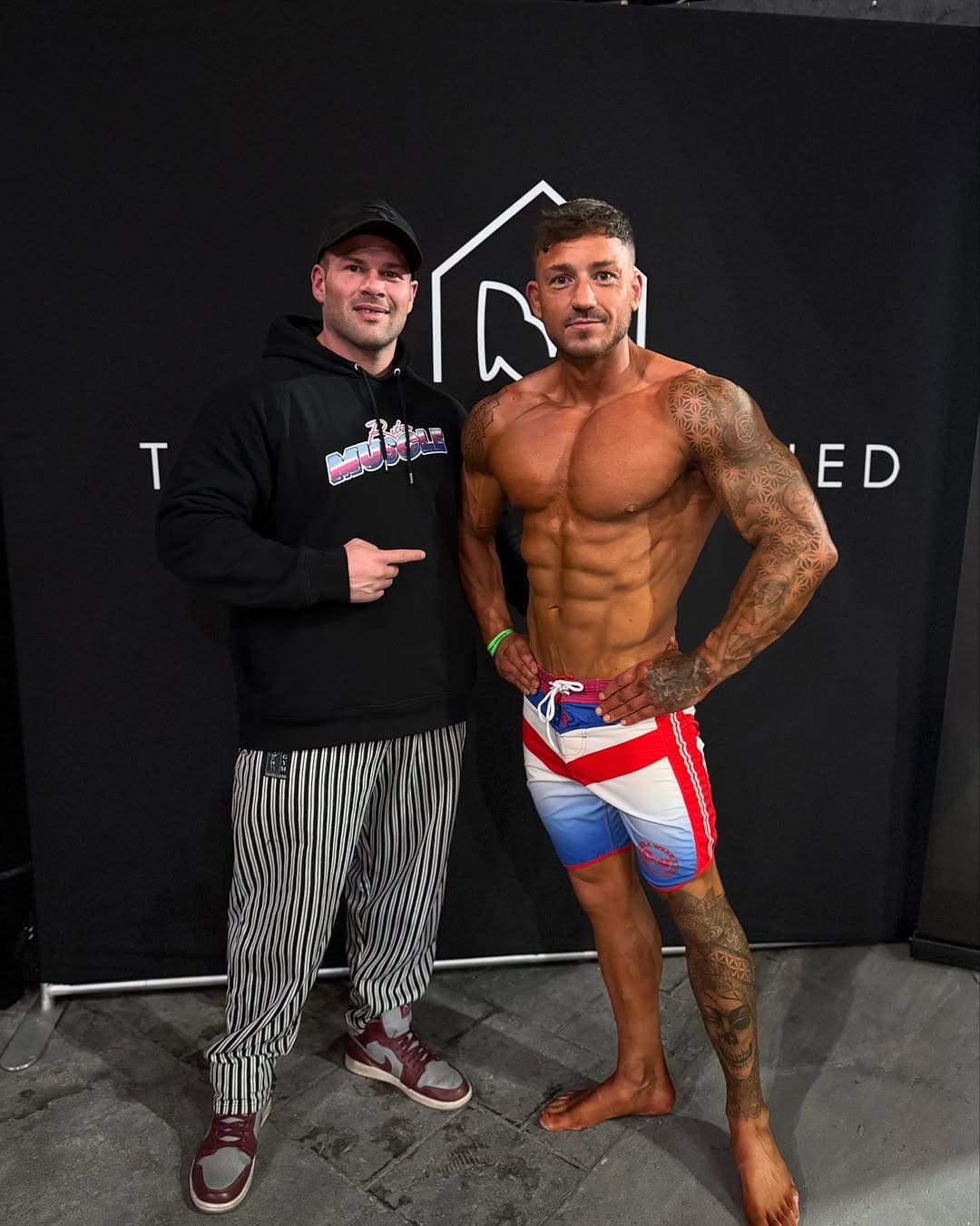 A ProFlow Physiques client in competition trunks standing with coach Joey Farrell against a dark backdrop at a show, competitor number still on the trunks.