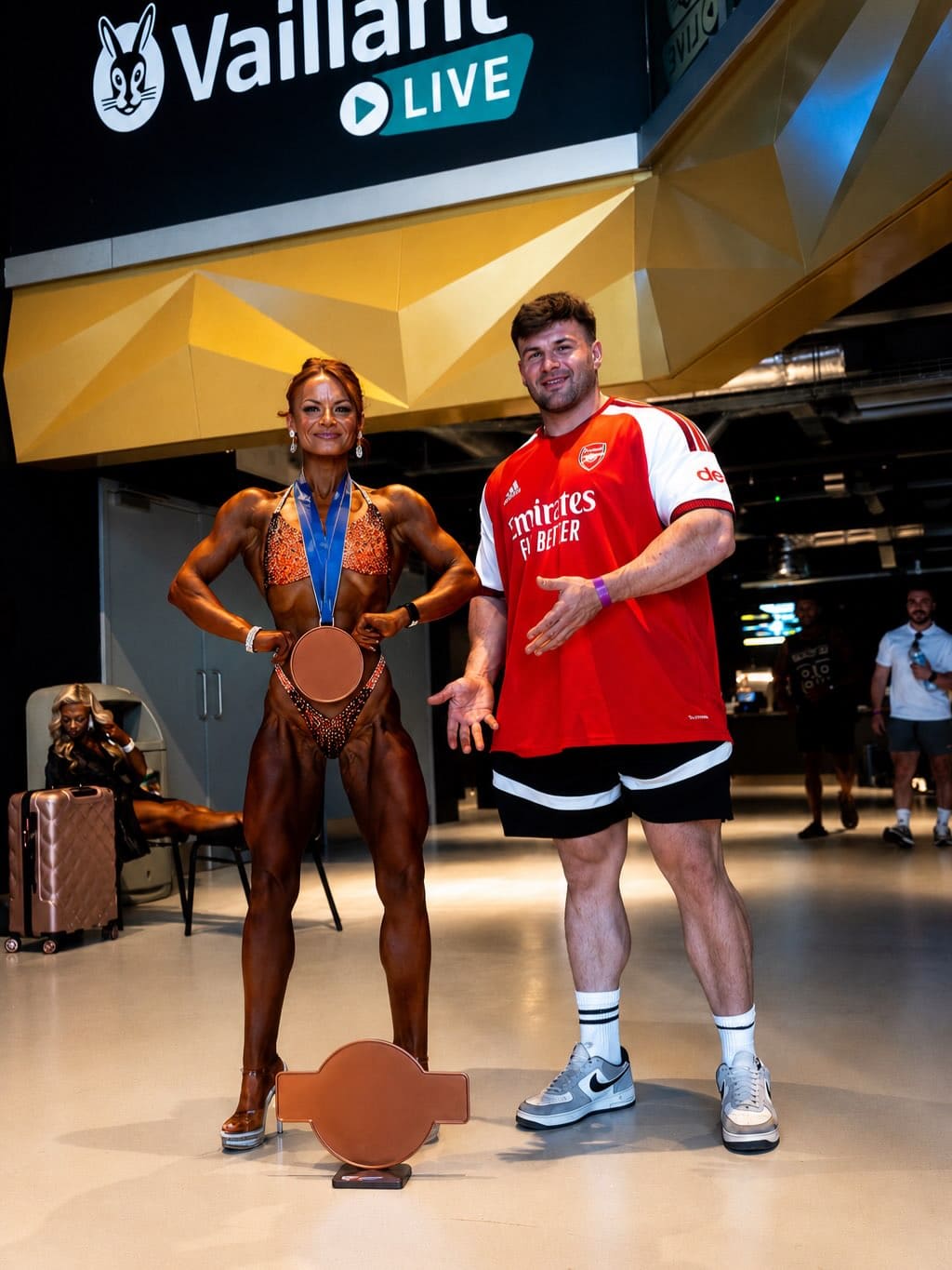 A ProFlow Physiques client in a competition bikini, medal round the neck, standing with coach Joey Farrell in the foyer of a competition venue.