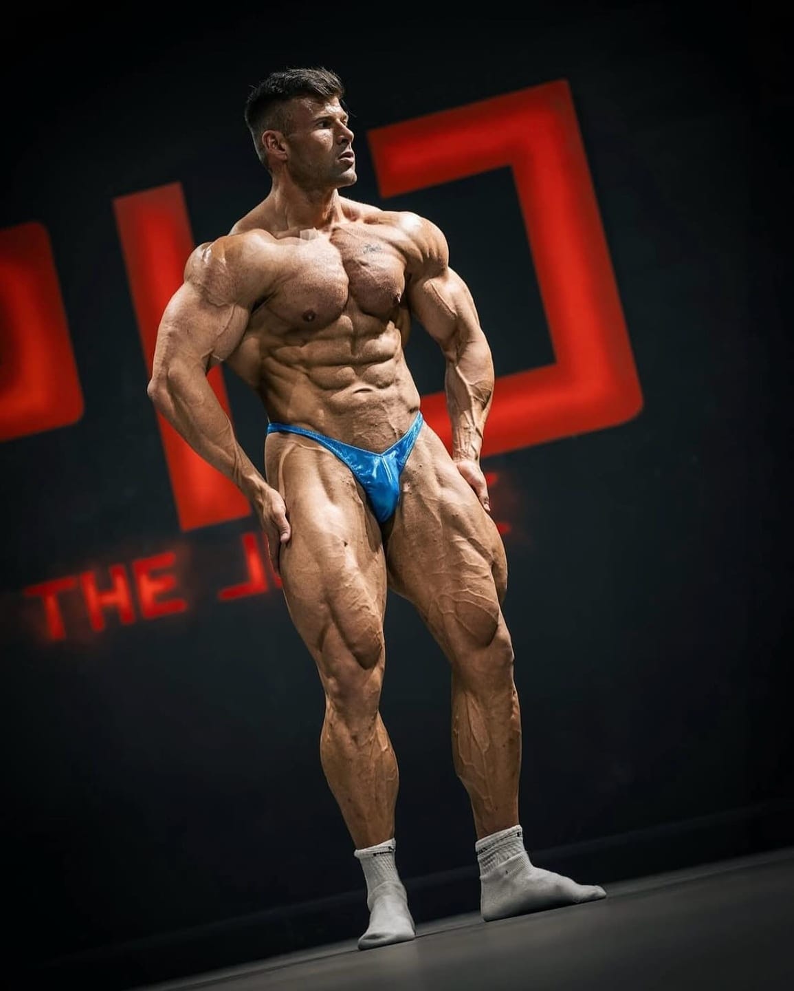 Joey Farrell, IFBB Pro and founder of ProFlow Physiques, posing under hard stage lighting against a dark backdrop.