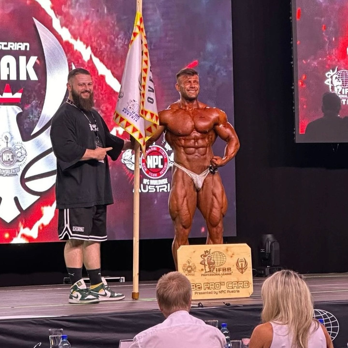 Joey Farrell on stage in contest condition, standing beside the plaque as he earns his IFBB pro card.