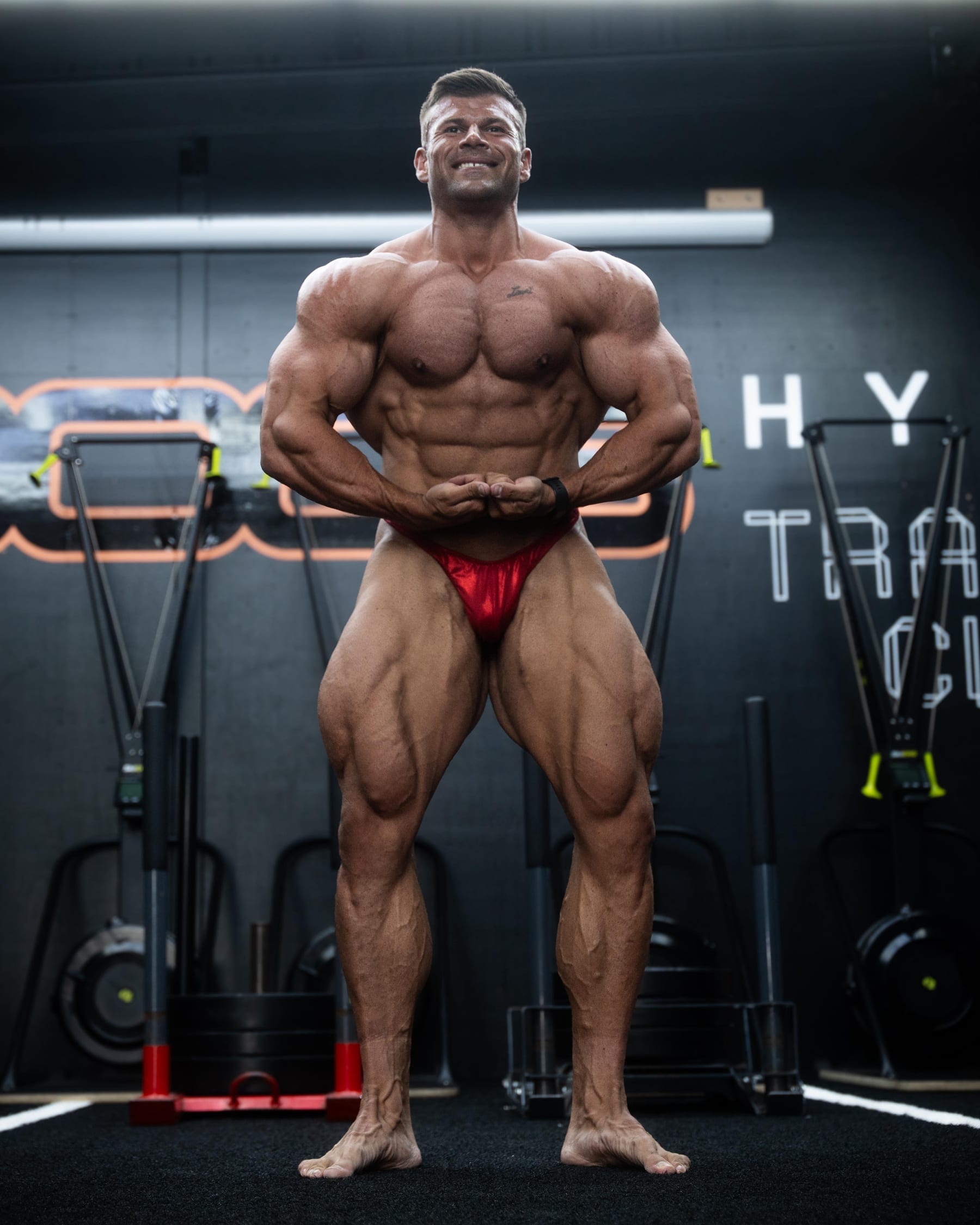 Joey Farrell in contest condition in a gym, holding a front pose and grinning.