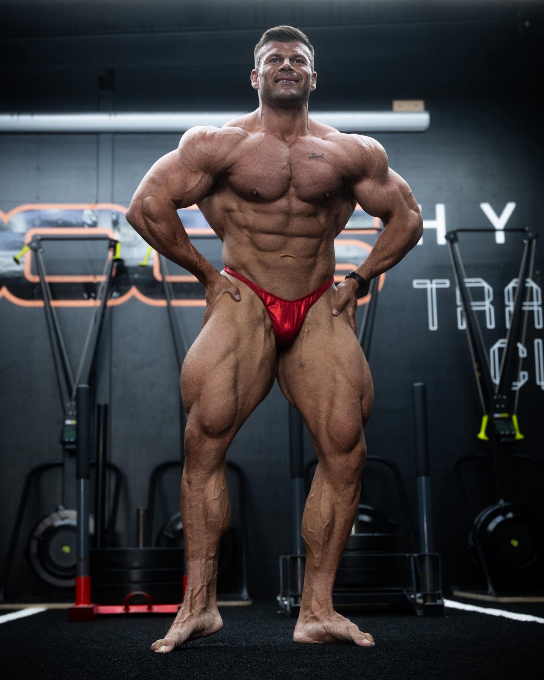 Joey Farrell in contest condition, standing relaxed and smiling in a gym under hard studio lighting.