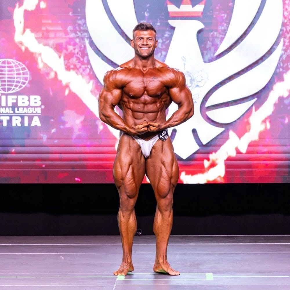 Joey Farrell on an IFBB stage in full contest condition, holding a front pose.
