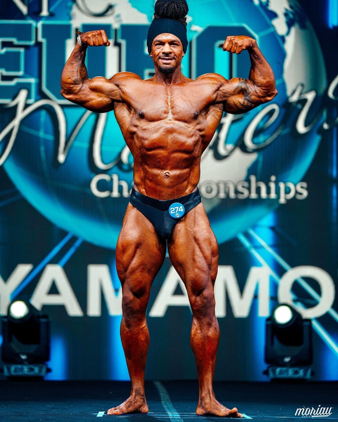 A ProFlow Physiques client alone on a competition stage under the lights, holding a front double biceps pose in front of a championship backdrop.
